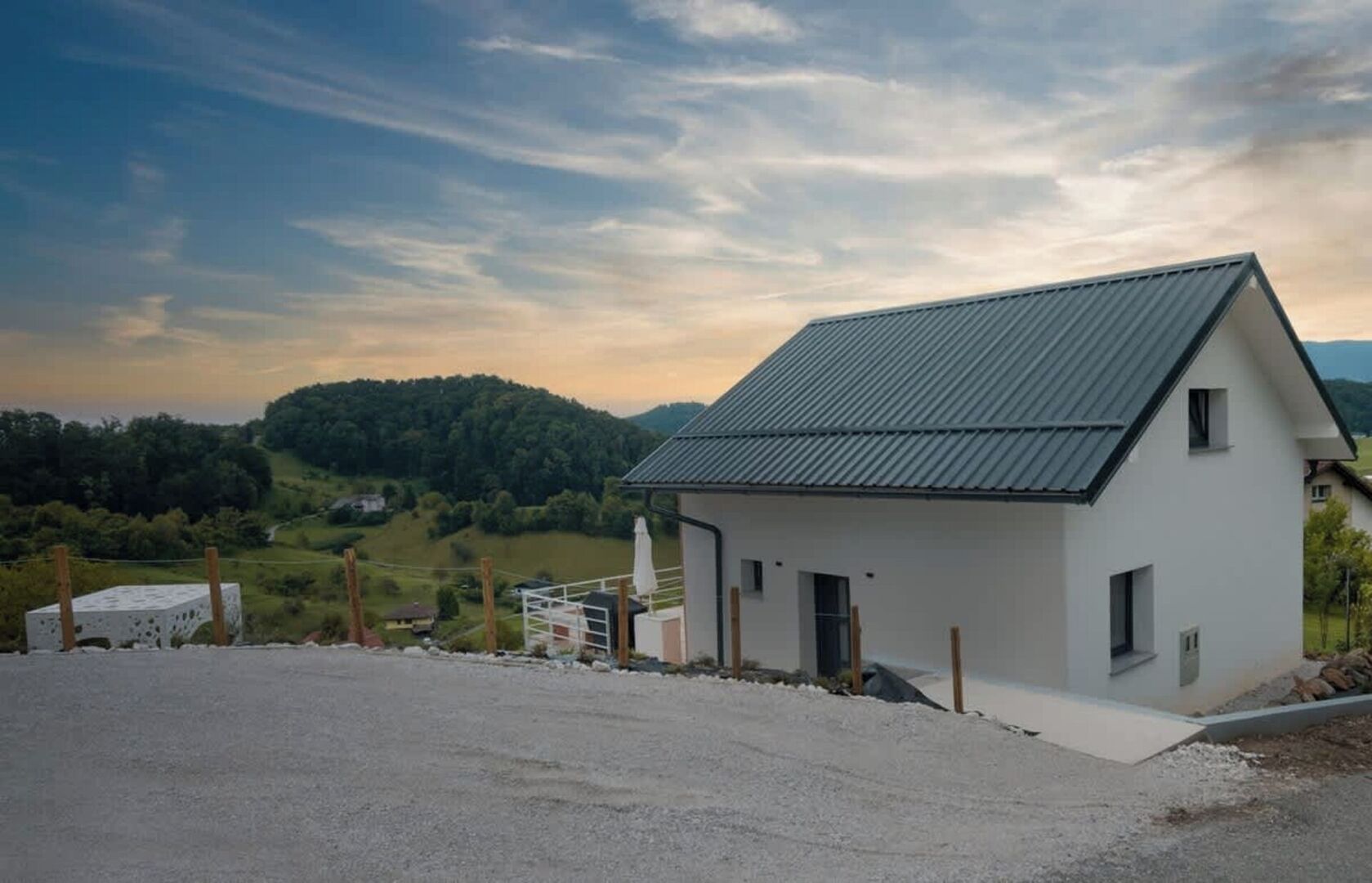 Un edificio blanco con techo inclinado, camino de grava y vista a un paisaje verde con colinas al fondo.