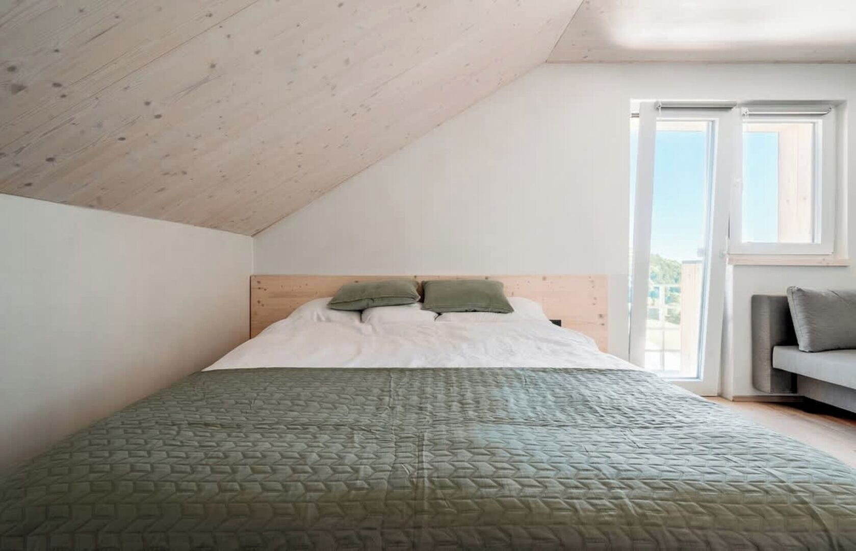 A bedroom with a sloped ceiling, a bed with a green quilt, a wooden headboard, and a grey sofa.