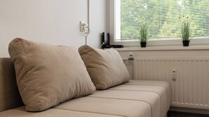 A beige sofa with two cushions, a radiator, and two potted plants on a windowsill.
