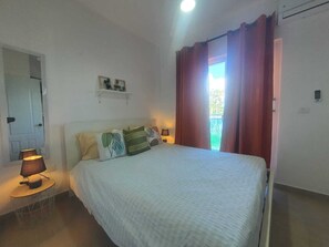 Ein Schlafzimmer mit einem Bett, Nachttischlampen, einem Spiegel, einem Regal mit Dekoration und Blick durch ein mit Vorhängen drapiertes Fenster.