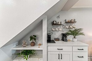Eine Küche mit weißen Schränken, einer Kaffeemaschine, einem Regal mit Tassen und einem Blumentopf.