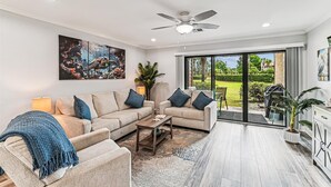 Un salon moderne comprenant un ensemble de canapés, une table basse, un ventilateur de plafond et une grande fenêtre donnant sur un jardin.