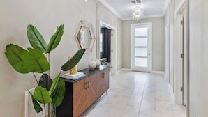 Un couloir avec un meuble en bois, de grandes plantes vertes, un miroir aux motifs géométriques et un lustre.