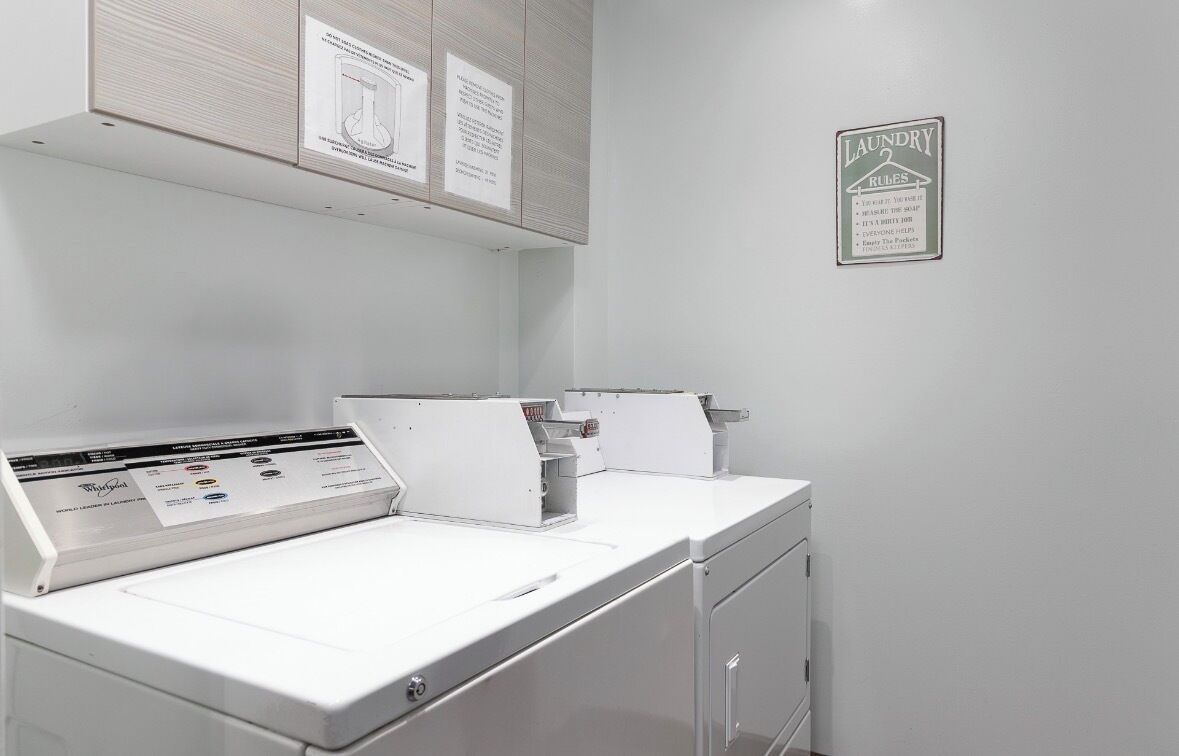 A laundry room with a white washing machine, a dryer, and a wall-mounted ironing board. There are instructional posters on the wall and a laundry rules sign.