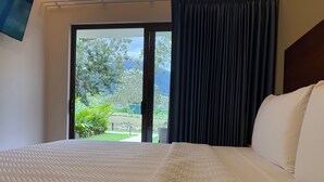 A hotel room with a bed, a view of greenery and mountains through a sliding door, and a dark curtain. - Arenal Volcanic Villas (La Fortuna)