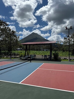 Uma quadra de tênis ao ar livre com um pavilhão, uma montanha ao fundo e vegetação ao redor.