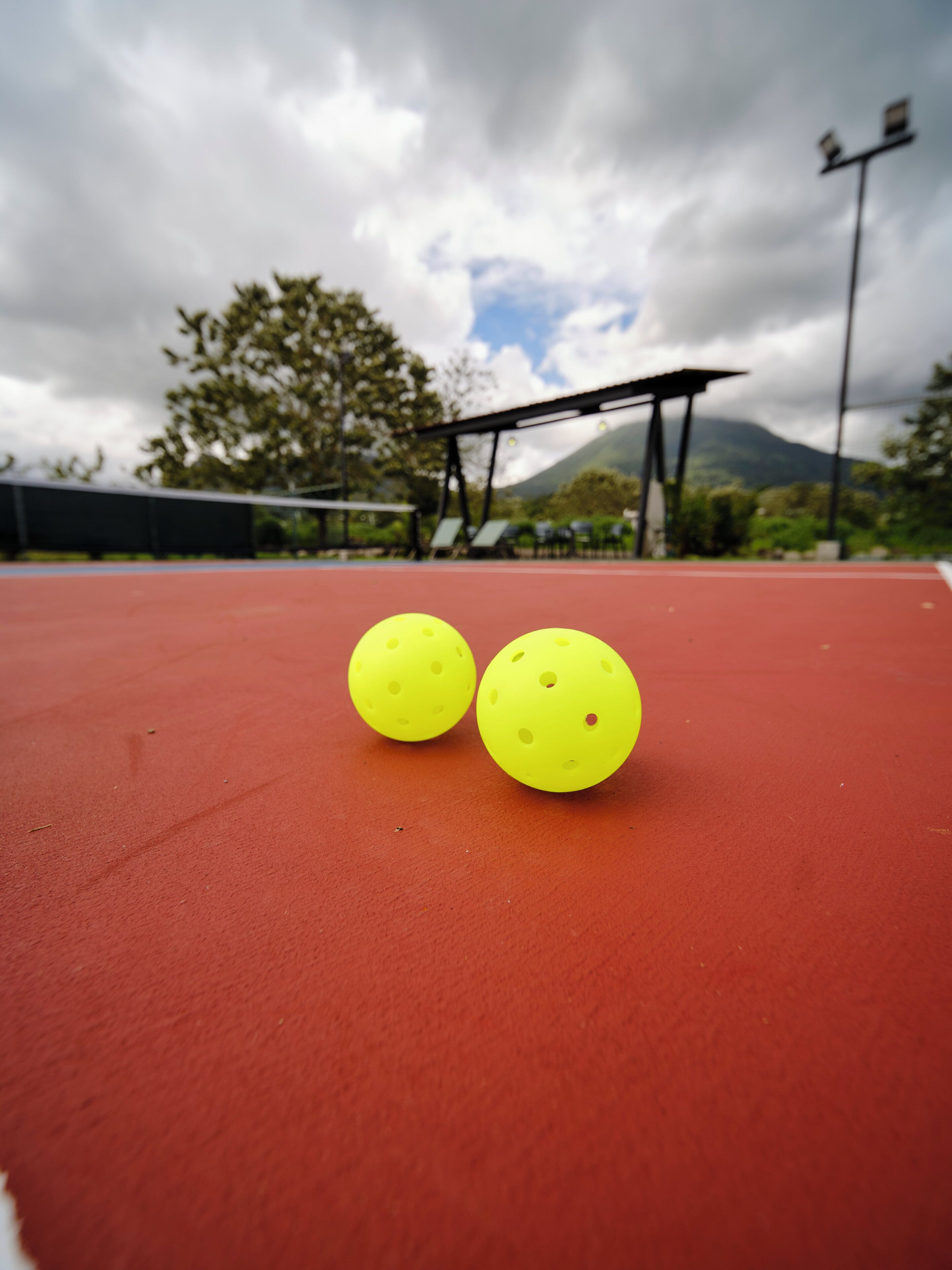 Two yellow tennis balls on a red court with a cloudy sky and a mountain in the background.
