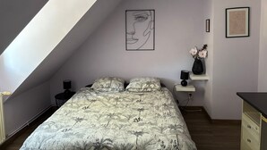 Et soveværelse med skrå vægge, en seng med dynebetræk med blomstermønster, et væghængt abstrakt maleri, en vase med blomster og et lille natbord med en lampe.