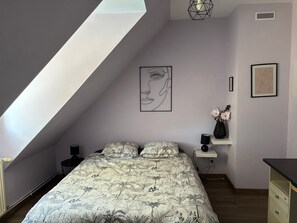 Ein Schlafzimmer mit schrägem Dach, einem Bett mit blumigem Überwurf, einem an der Wand hängenden abstrakten Gemälde, einer Vase mit Blumen und einem kleinen Nachttisch mit Lampe.