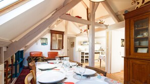 Un coin repas avec une table dressée pour quatre personnes, des chaises en bois, une moquette rouge et une vue sur une autre pièce où se trouve une cuisine.
