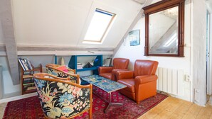 Une pièce de style « loft » avec un plafond en pente, une lucarne, un grand miroir, un tapis rouge à motifs, une chaise en bois, un fauteuil en cuir et une bibliothèque.