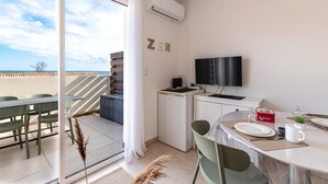 Une chambre moderne dotée d’une porte-fenêtre coulissante donnant sur un balcon, d’un téléviseur à écran plat, d’une petite kitchenette et d’un coin repas avec une table mise pour deux personnes.