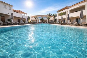 Une piscine aux eaux cristallines, bordée de chaises longues et d’ombrelles, et entourée d’immeubles dotés de balcons.
