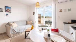 Un salon moderne comprenant un canapé, une table à manger dressée avec des assiettes, des tasses et un vase, ainsi qu’un coin cuisine doté d’un plan de travail et de placards.