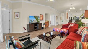 Ein geräumiges Wohnzimmer mit einer großen Couch, einem Couchtisch und einem Fernseher. Es gibt einen Deckenventilator, einen Essbereich mit Tisch und Stühlen sowie eine Küche im Hintergrund.