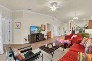 A spacious living room with a large sofa, a coffee table, and a television. There is a ceiling fan, a dining area with a table and chairs, and a kitchen in the background. - Treviso Bay Retreat I Resort-Style Amenities I Relaxing Lakeview Escape (Naples)