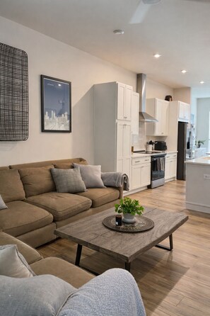 A modern living room with a sofa, a coffee table with a plant, and a kitchen area with white cabinets and appliances. - FIFA 2026 RETREAT - 2BR Sleeps 6 - Bikes - BeltLine to Stadium - Walkable (Atlanta)