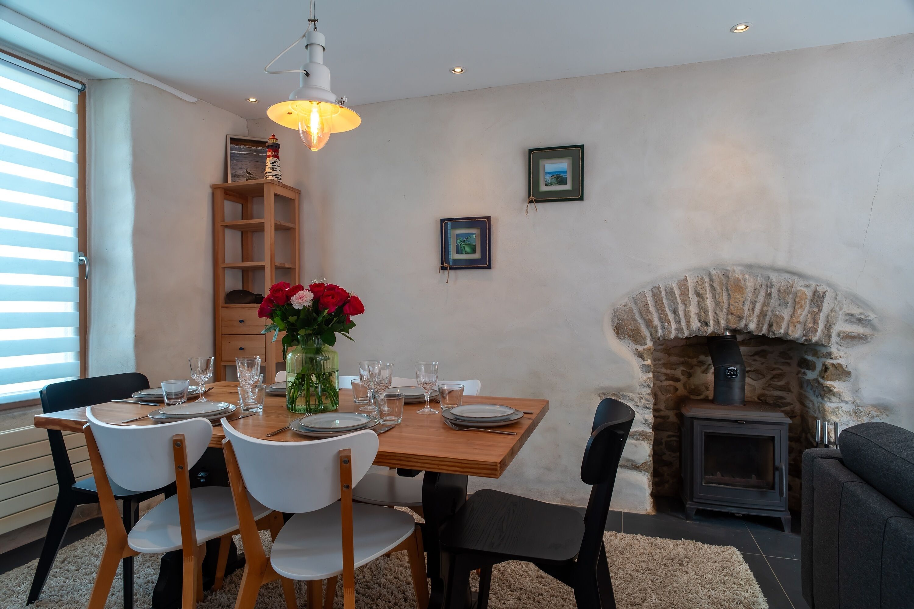 Un coin repas avec une table dressée pour quatre personnes, une cheminée en pierre et une étagère en bois ornée d’un vase rempli de fleurs rouges.