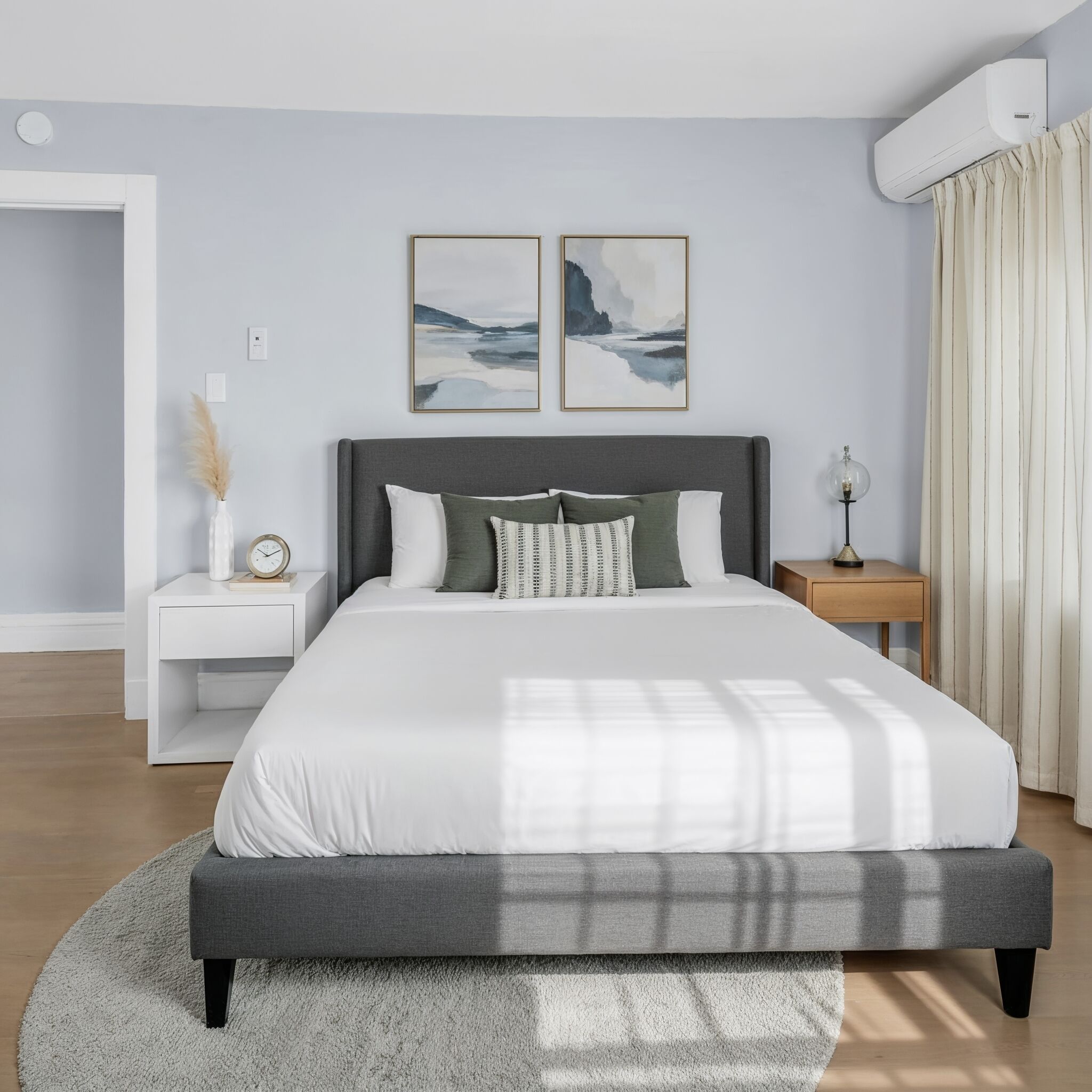 Une chambre moderne avec un grand lit, des tables de chevet, une petite table de nuit, un climatiseur fixé au mur et deux tableaux encadrés au-dessus du lit.