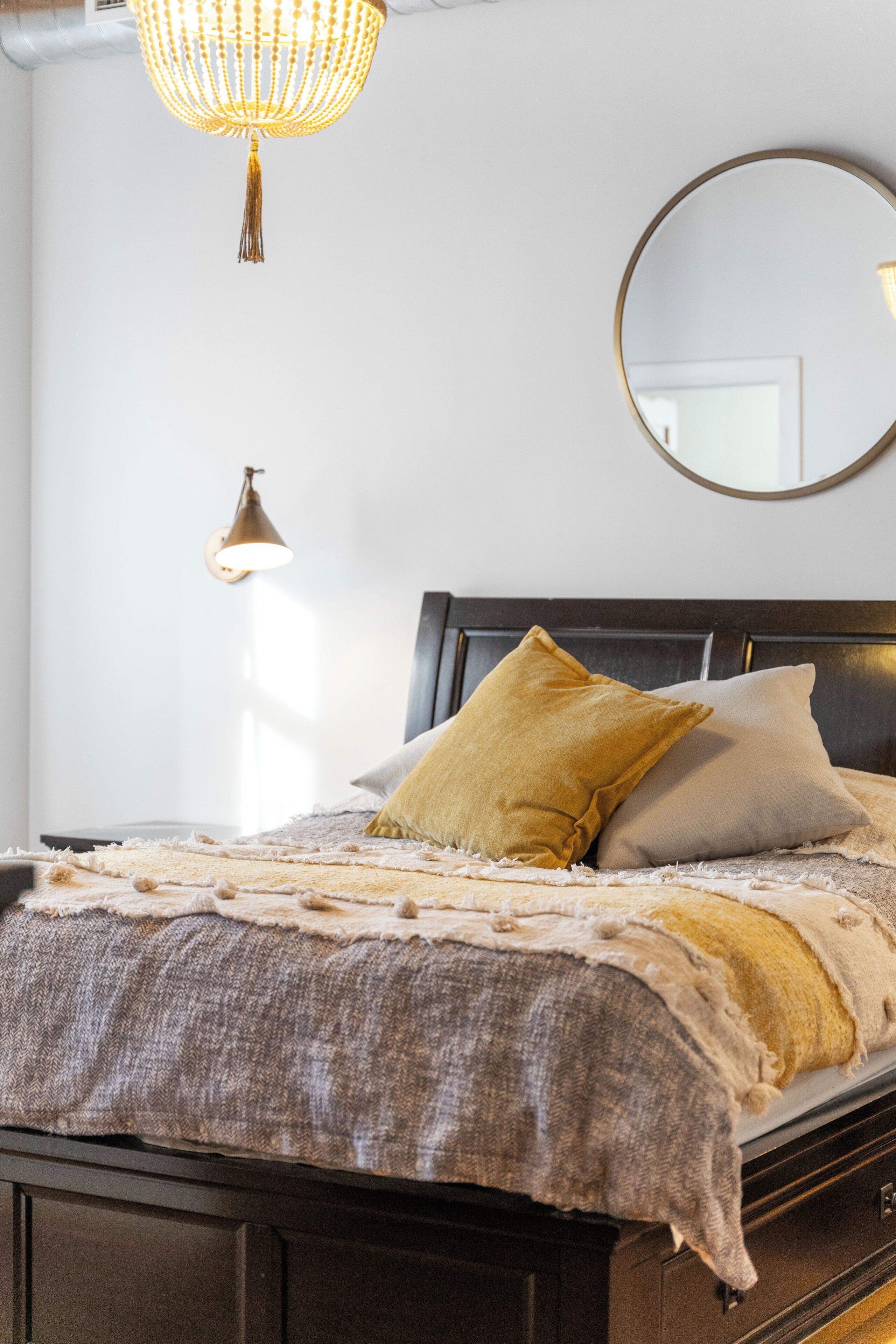 A bed with a dark headboard, a round mirror on the wall, and a hanging light fixture.