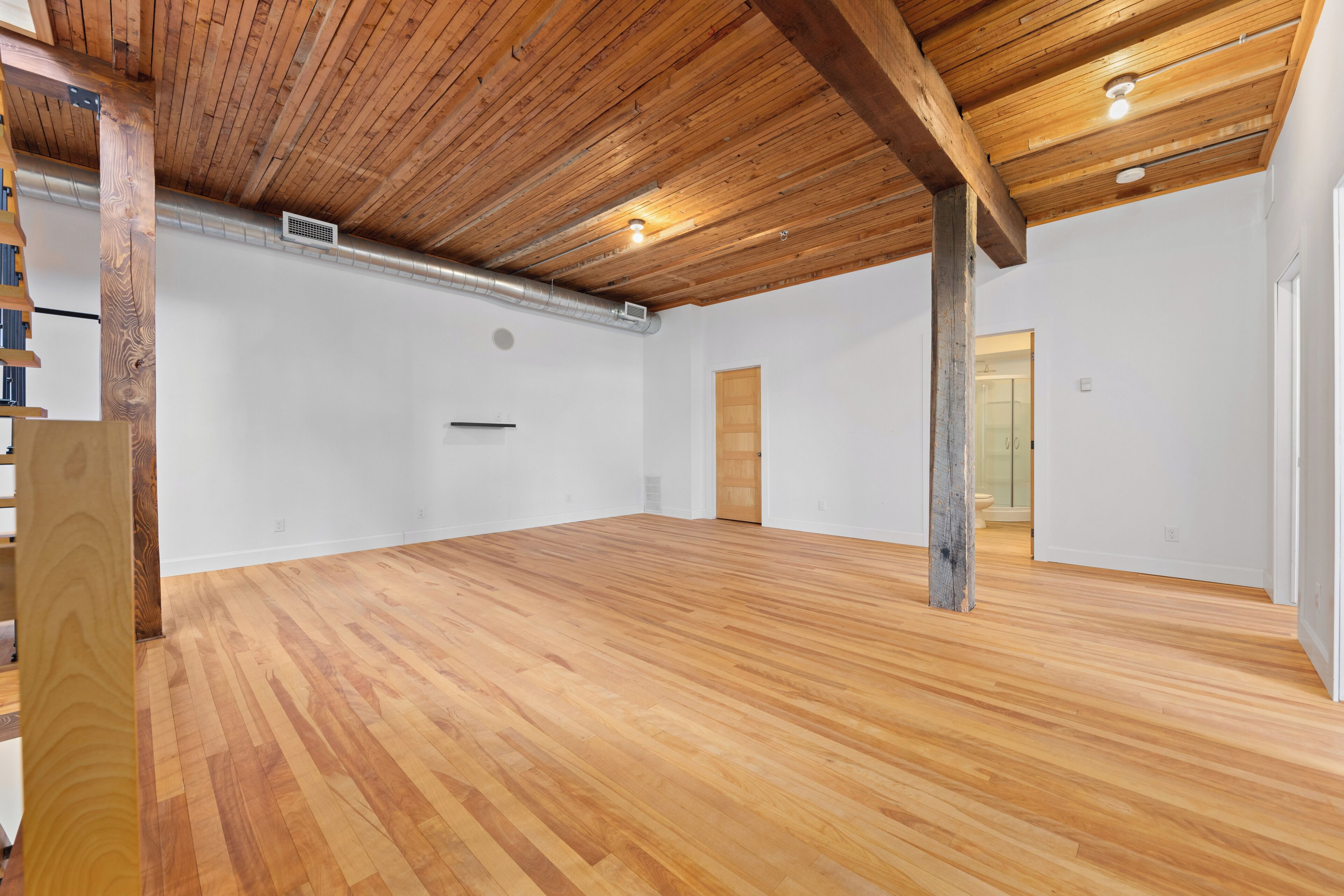 Una stanza spaziosa con pavimento in legno, travi a vista in legno, pareti bianche e una porta che dà su un'altra stanza.