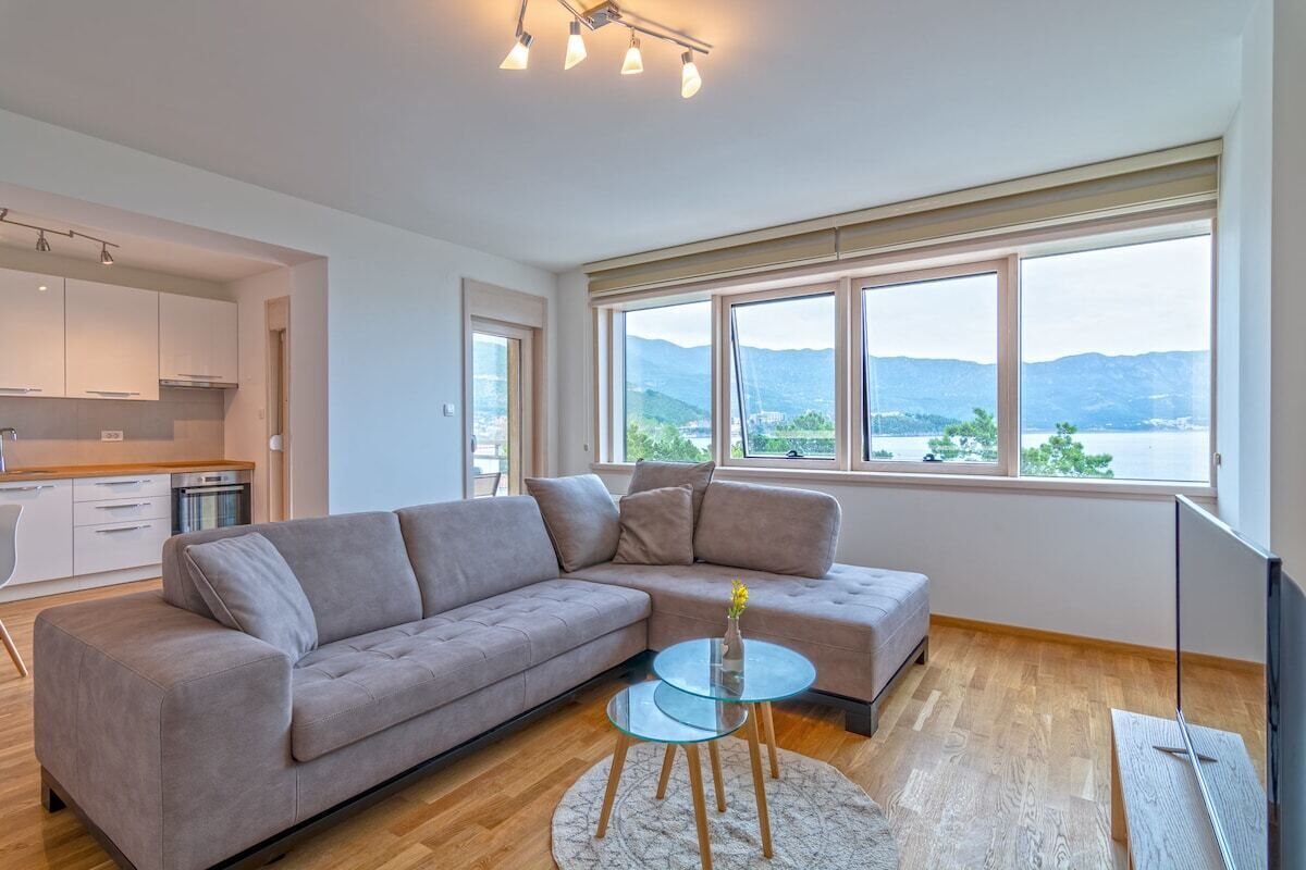 A modern living room with a sofa, a glass coffee table, and a view of mountains through large windows.