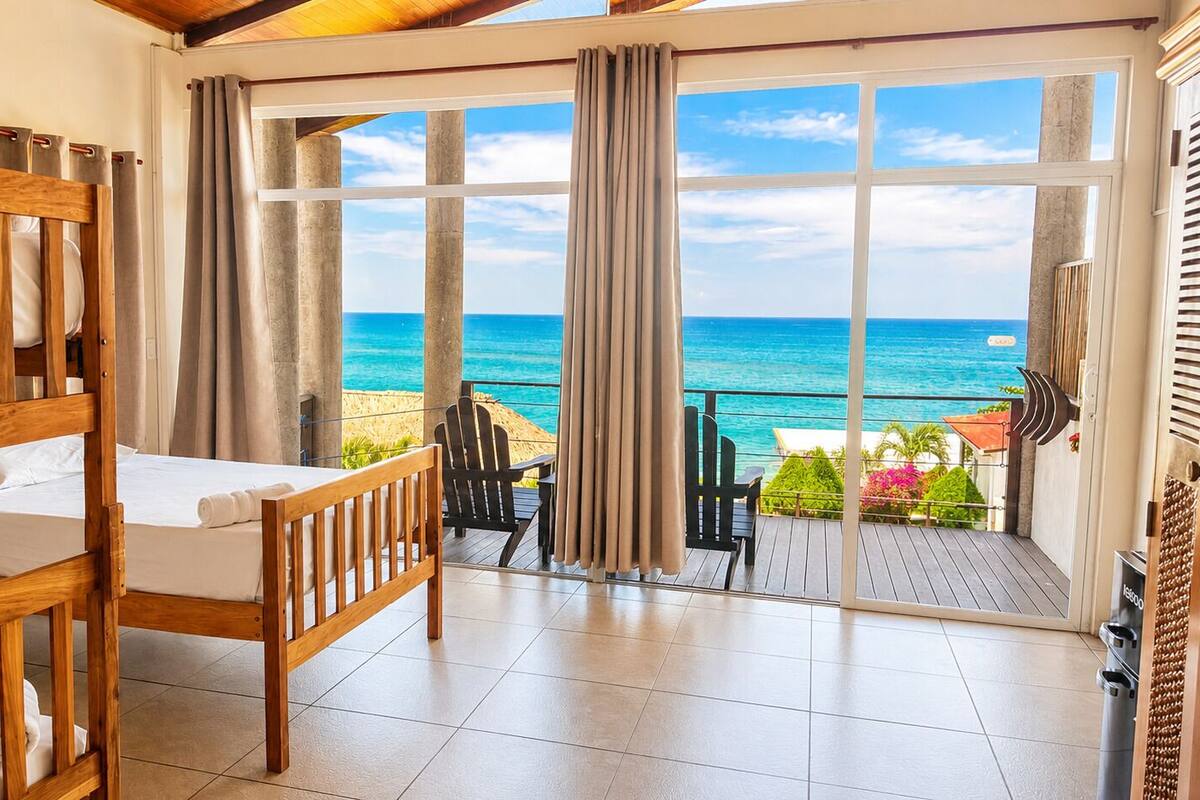 Une chambre avec un lit en bois, un balcon donnant sur l’océan et un coin salon avec des chaises et une table.