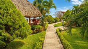 A pathway leading to a thatched-roof structure surrounded by lush greenery and palm trees. - LAOLA SURF RESIDENCE (El Sunzal)