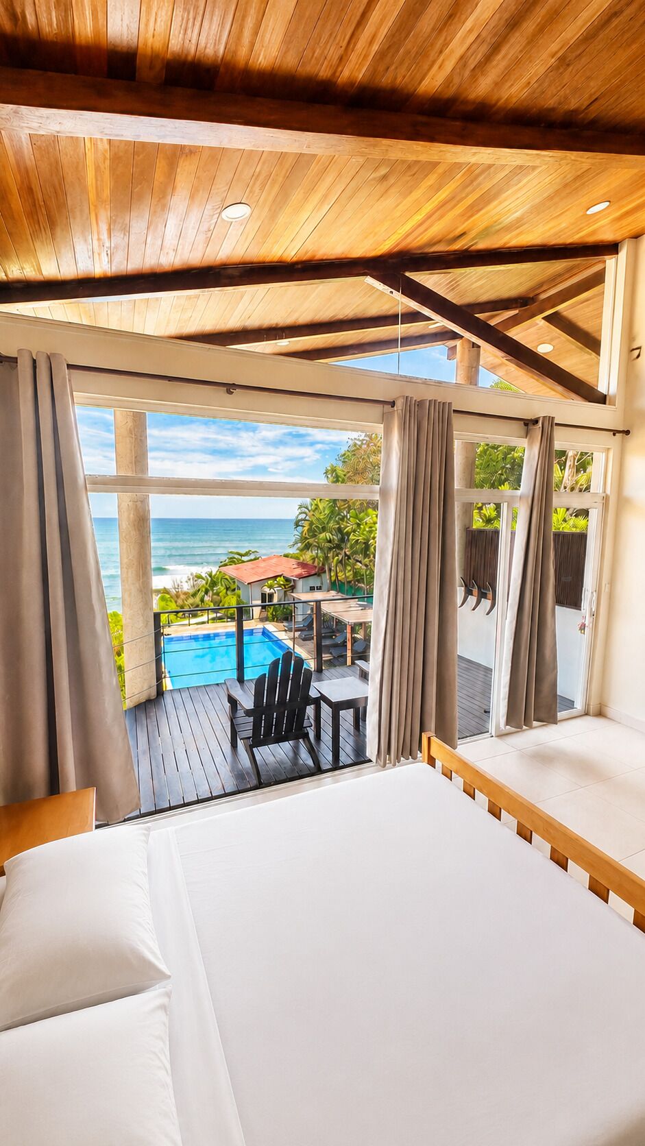 Une chambre avec un plafond en bois, un grand lit et une vue sur une piscine extérieure et un paysage tropical.