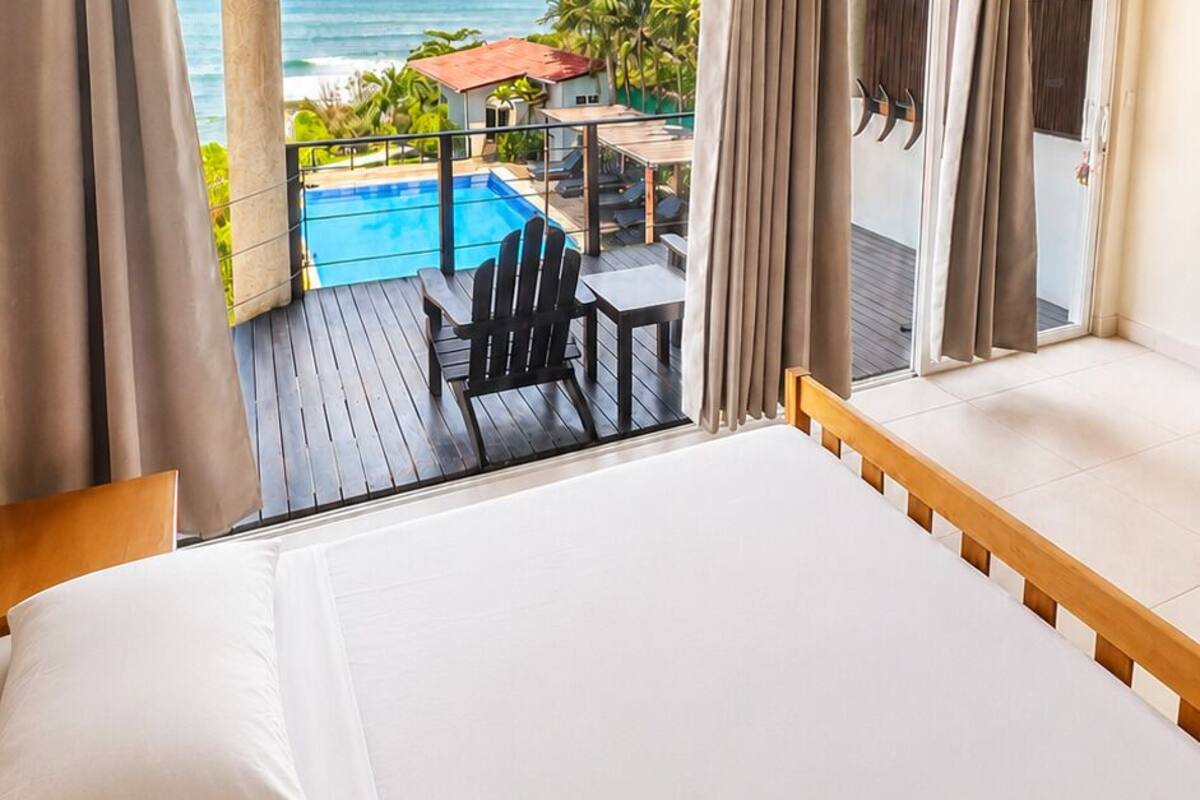 Une chambre avec un plafond en bois, un grand lit et une vue sur une piscine extérieure et un paysage tropical.
