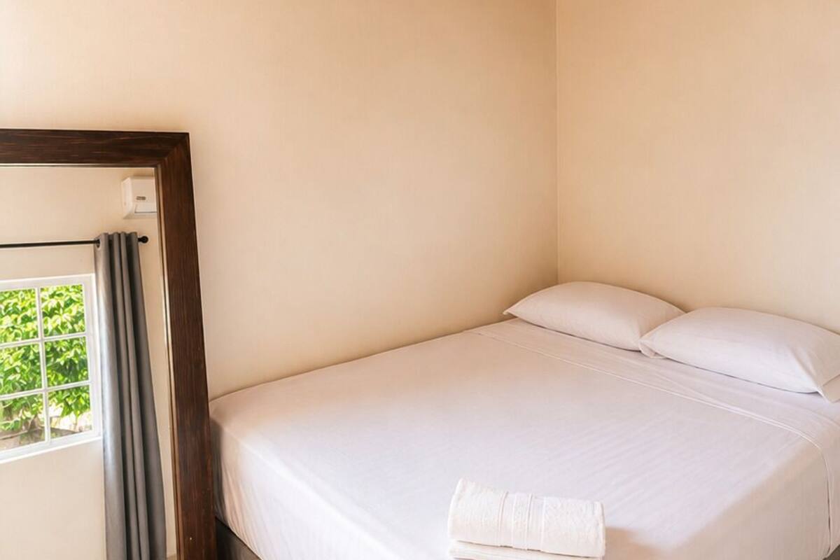 Une chambre avec un plafond en bois, un lit recouvert de draps blancs, une fenêtre avec des rideaux et une petite table de chevet.