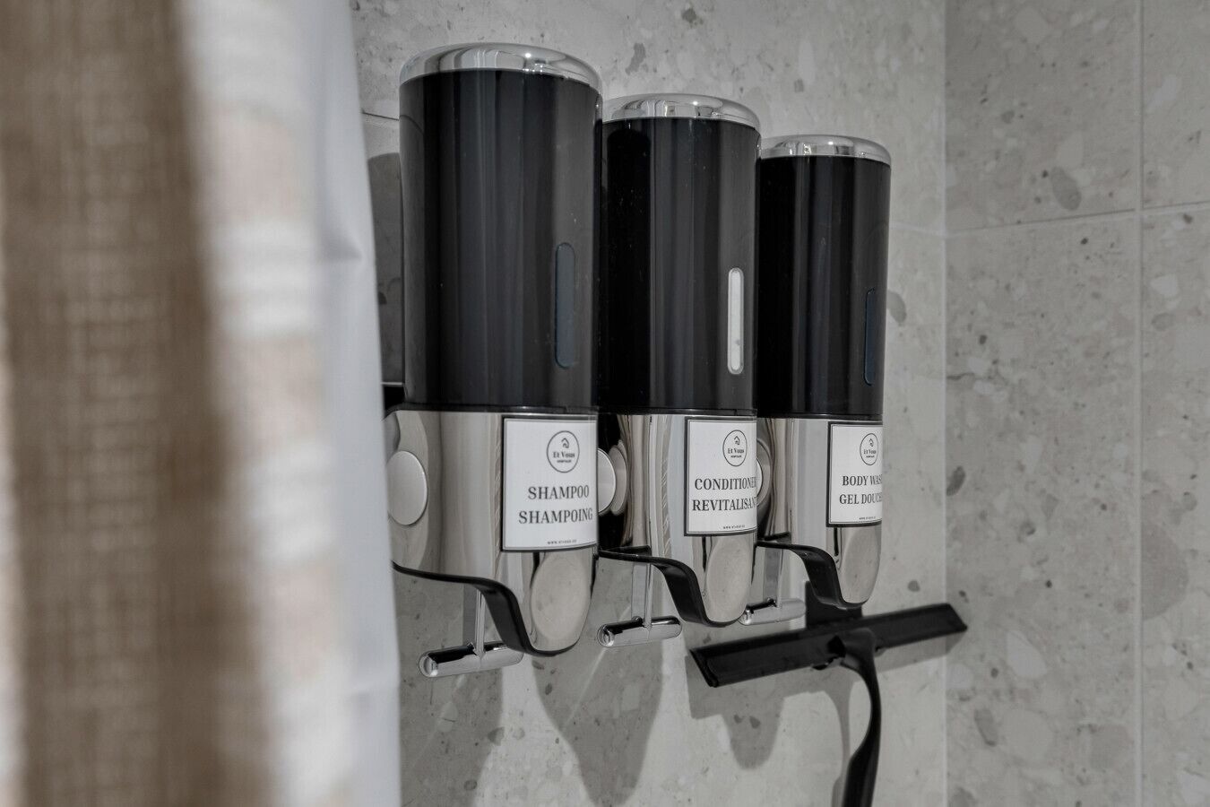Seinään kiinnitettävä annostelulaite, jossa on kolme pulloa: yksi merkitty nimellä ”Shampoo”, toinen ”Hoitava ja elvyttävä hiustenhoitoaine” ja kolmas ”Hierontageeli”.