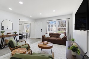 Ein modernes Wohnzimmer mit einem braunen Sofa, grünen Sesseln, einem runden Holztisch und einem Essbereich mit weißem Tisch und Stühlen.