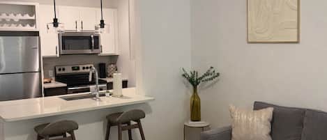 Ett modernt kök med en central köksö, barstolar, ett matbord, en takfläkt och en grå soffa i ett vardagsrum.