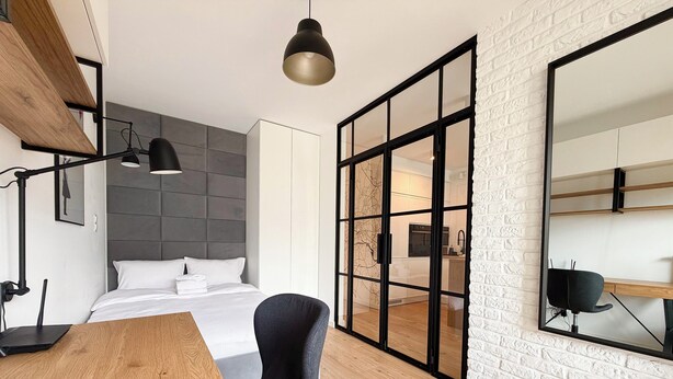 A modern hotel room with a bed, a desk, a chair, a large mirror, and a sliding glass door leading to a kitchen area. - Street Art Loft Apartment (Wrocław)