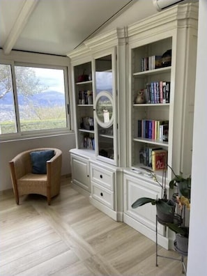 Ein Zimmer mit großem Fenster, einem Korbstuhl, einer weißen, fest installierten Bücherwand und einem Blumentopf.