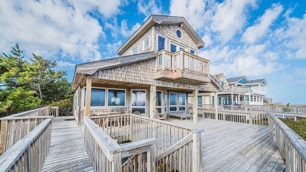 Et træhus ved stranden med terrasse, store vinduer og en balkon.