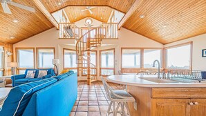 En rummelig opholdsstue med en blå sofa, et køkken med en stor ø, og en spiraltrappe der fører op til en etage over.