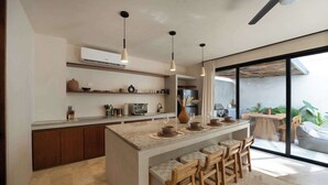 Una cocina moderna con una isla central, taburetes de bar, iluminación colgante y un amplio ventanal corredero que da a un área exterior.