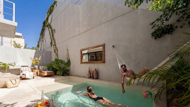Piscina com uma pessoa boiando, outra pessoa pendurada em uma rede, e área de descanso com espreguiçadeiras e uma mesa com frutas e bebidas.
