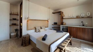 Quarto com cama grande, mesas de cabeceira, um banco de madeira, um banco embutido na frente da cama, luminária de cabeceira, prateleira com objetos decorativos e uma pequena área de cozinha.