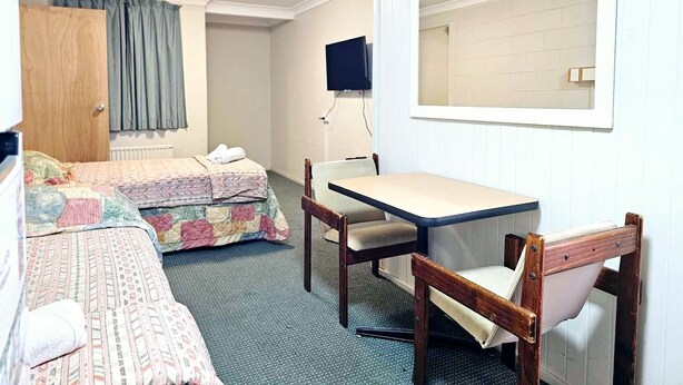 Een kleine, goed verlichte kamer met een bed, een bureau en een stoel, met een televisie en een raam.