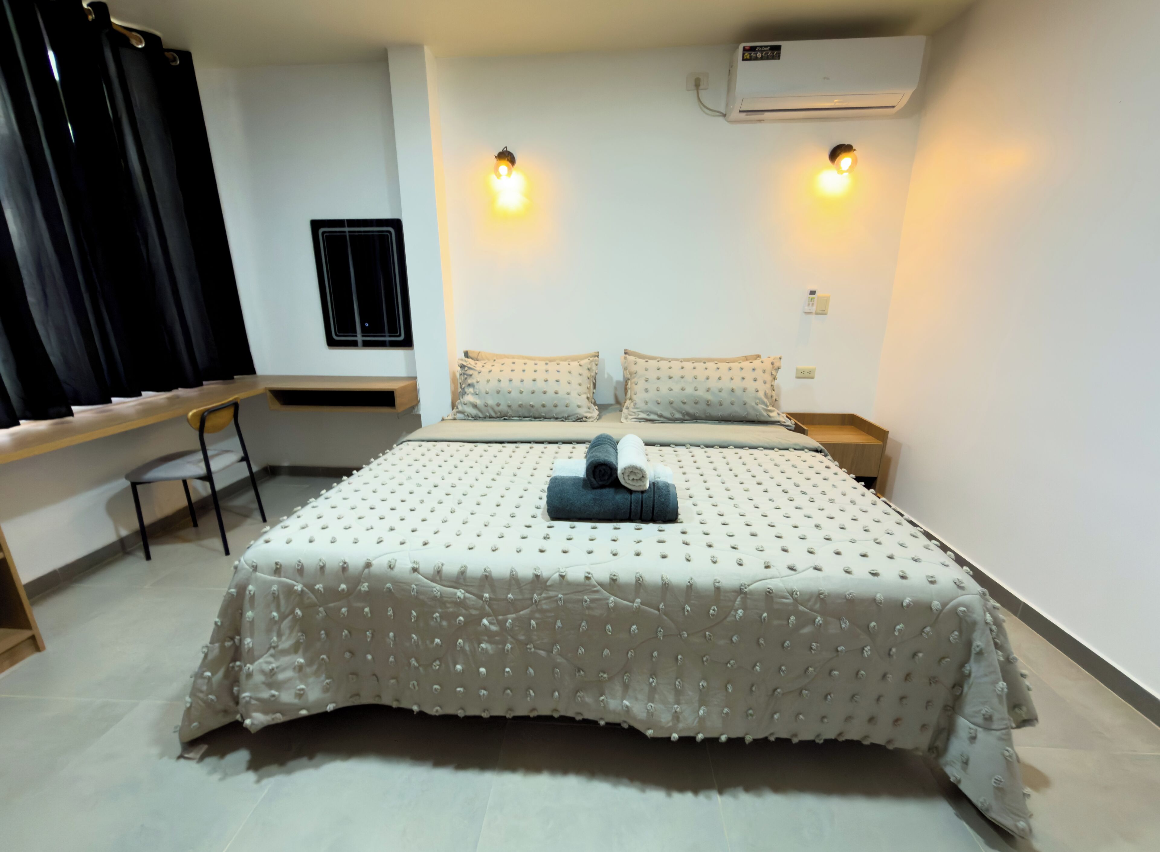 A neatly arranged bedroom with a bed, pillows, a bedside table, a desk, and an air conditioning unit.