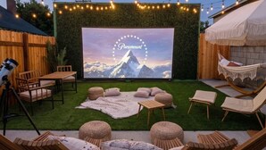 Une terrasse extérieure dotée d’un écran de projection diffusant des images de « Paramount », entourée de meubles en bois et illuminée de guirlandes lumineuses.