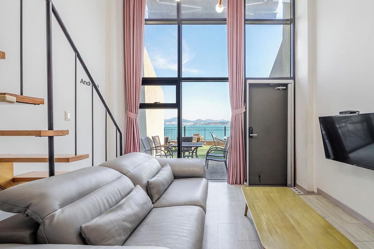 A modern living room with a sofa, a TV, a staircase, and a view of the sea.