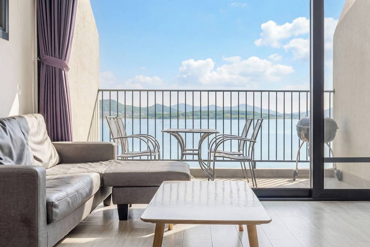 A balcony with a sofa, chairs, and a table, overlooking a body of water and hills.