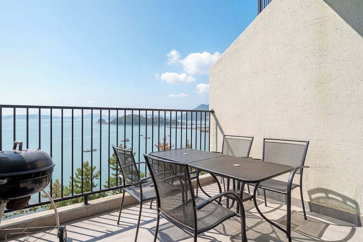 A balcony with a grill, table, and chairs overlooking a body of water and distant land.