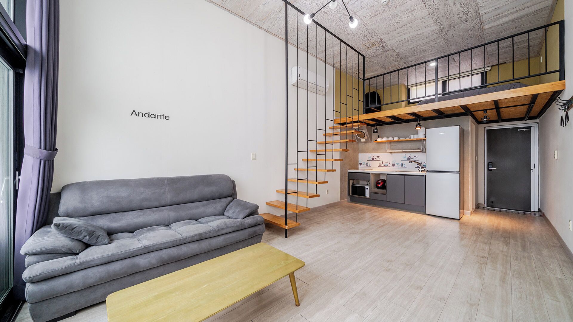 Un espacio habitable moderno con un sofá gris, un banco de madera, una escalera con escalones de madera, una zona de cocina y un altillo con suelo de madera.