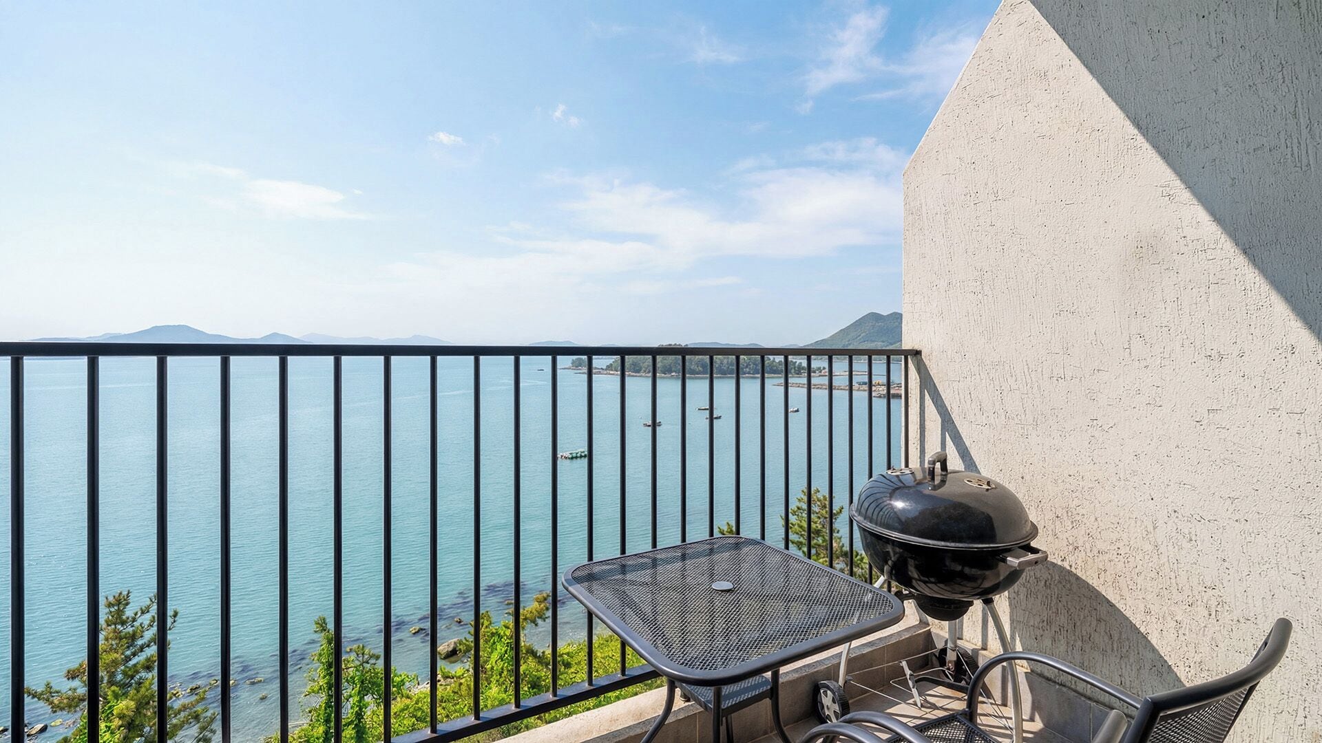 A balcony with a view of a body of water, a grill, and a table.
