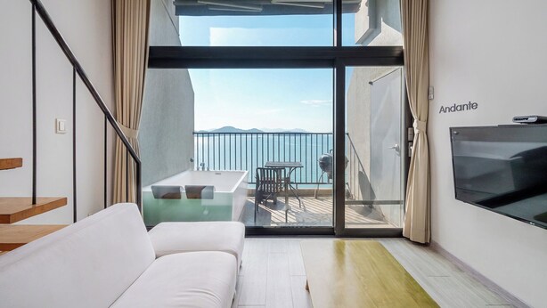 A modern living room with a sofa, a TV, and a balcony with a table and chairs. - Yeosu Andante Spa Pension (Yeosu)